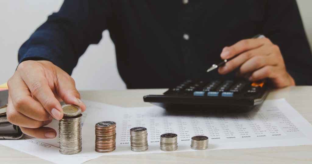 A pile of coins, an adult behind them about to pick up a coin while the other hand rests above a calculator