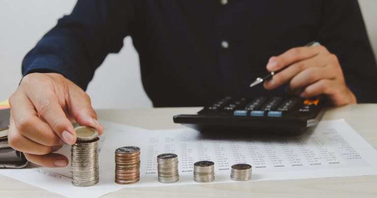A pile of coins, an adult behind them about to pick up a coin while the other hand rests above a calculator