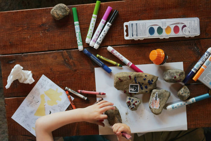 A child's hands over a tabletop scattered with rocks, paper, paints and pens