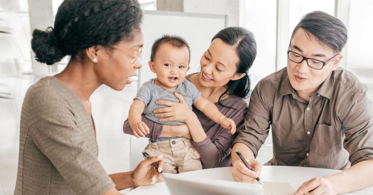 3 adults and a baby sit around a table while one adult signs or writes something on the table