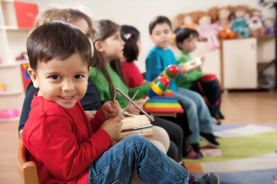 Nursery leadership and governance Image - A toddler looks at the camera while holding a musical triangle and other bandmates are sat nearby